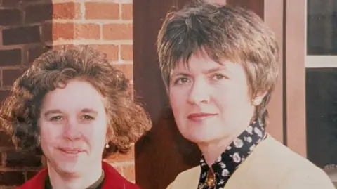 Hope House An old photograph of two women. Barbara on the left has short curly brown hair and a red jacket. The woman on the right has a short brown pixie haircut and is wearing a black top with white flowers and a beige suit jacket