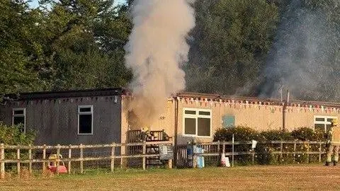 A single-storey building decorated with bunting with smoke pouring from an open door as firefighters enter