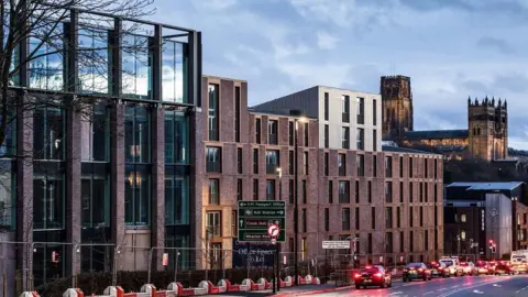 Milburngate. The development is several storeys high and has large glass windows. Durham Cathedral can be seen in the background and a number of vehicles are being driven along the main road in front of the development.