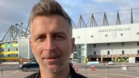 BBC A head-and-shoulders image of a man with grey stubble and grey hair in a quiff. He is standing outside, with Norwich City's Carrow Road stadium behind him 