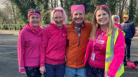 Three women and a man pose in a line and smile while standing in a large car park. The women are wearing pink tops and pink headscarf bows. The man is wearing an orange zip-up top, blue jeans and a pink headscarf. 