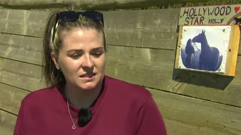 A woman with light brown hair. The woman is white, she is wearing a red shirt and has sunglasses on her head. She is visibly sad. She is sat in front of a wooden background. To the right of her is a black and white picture of her kissing a horse. Above the photo is a plaque which reads Hollywood Star Holly