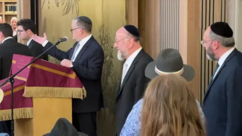 The Chief Rabbi walks into the synagogue as attendees of a service look on, including a man behind a stand.