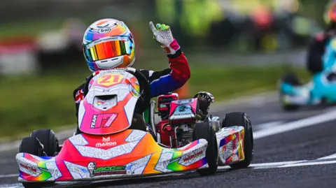 Josh East Photography A motorsport racer in a small multi-coloured kart wearing a colourful helmet is pictured on a racecourse with his left hand in the air. 
