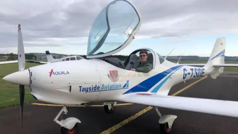 The 16 year old pilot Sam, wearing sunglasses and a green flying suit, is sitting inside the cockpit of a small white two seater plane by the side of the runway.