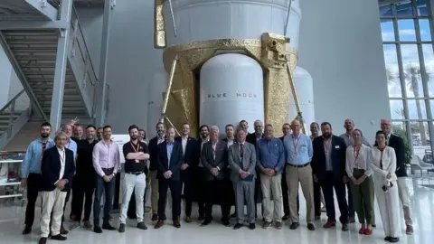 A delegation of space industry representatives standing in front of a Blue Moon lunar lander. The large craft is white with sections painted gold.
