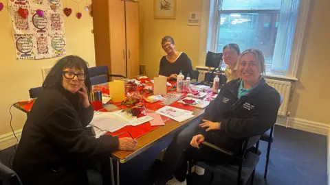 A yellow room with a large table in the middle. On the table are four women posing to the camera. The one closest to the camera has her eyes closed and black glasses. The others are smiling. There is a window in the background. There are lots of crafts on the table as people are making cards. There are lots of glue bottles also. The table has red tablecloth. 