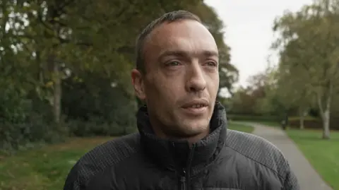 A man in a black coat stands on a path in a park, surrounded by green grass and trees. He has an inquisitive expression, looking slightly beyond the camera.