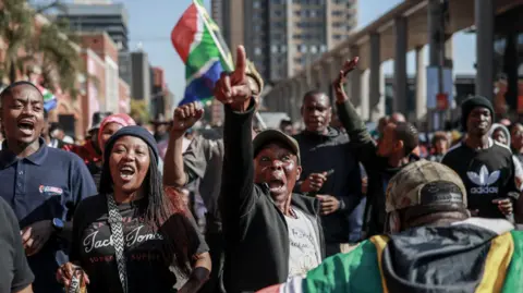 Protesters chant slogans as they march during an anti-migrant demonstration in Pretoria - 30 June 2026. One of the points into the air and behind him a South African flag can be seen. 