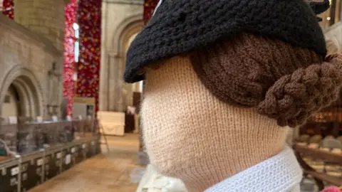 A close-up of the side view of a knitted female figure in World War Two dress in Peterborough Cathedral. It has a black hat, a cream face and brown hair in a bun. Beyond it are stone-flagged floors, cabinets with more knitted figures and stone arches. 