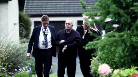 Firecrest Films A handcuffed 77-year-old man is led from his house by two police officers