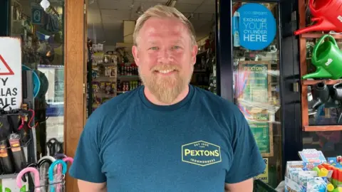 A head and shoulders shot of Bobby Paul, stood outside his business Pextons Hardware Store. Mr Pexton has short fair hair and a beard, and wears a blue t-shirt with the business logo on the chest.
