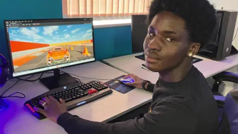 A boy sat at a computer monitor with an orange cartoon car on the screen. He is holding the computer mouse and is turned to look at the camera