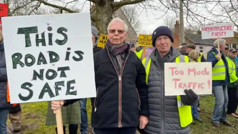 Villagers in Coltishall on a demonstration. Two men hold placards, with others in the backdrop. The placards read: "This road is not safe"; another reads: "Too much traffic"; and another says: "Infrastructure first".
