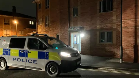 A police van parked on a residential street at night. The van is marked with blue and yellow panels and the word “POLICE”, and its headlights are switched on. Behind it is a red-brick apartment building with a lit entrance and several windows visible.

