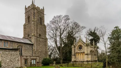 Geograph/Ian Capper A flint-built medieval church on the right and on the left is a Gothic-revival style mausoleum in cream stone, surrounded by black railings in a church yard. 