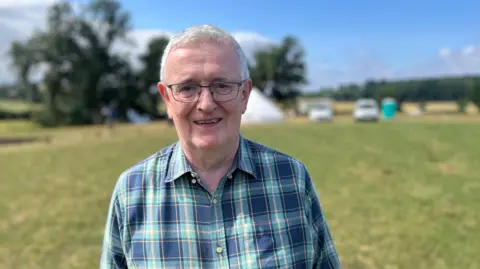 A grey-haired man with glasses, wearing a green and blue tartan shirt, looks at the camera