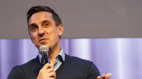 Former professional footballer Gary Neville with brown hair wearing a blue shirt and navy V-neck jumper. He is laughing as he talks into a microphone he is holding with his left hand. 