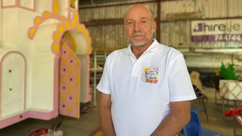 a man wearing a white 'Battle of Flowers' branded polo shirt stands with his hands together while smiling for the camera.