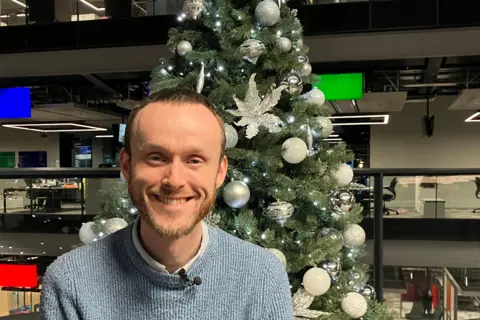 Food historian, Carwyn Graves is looking straight at the camera and he is smiling. He has short dark hair and a short ginger beard. He is wearing a blue knitted jumper with a shirt collar sticking out. The picture is festive as Carwyn is sat in front of a Christmas tree with silver decorations on it.