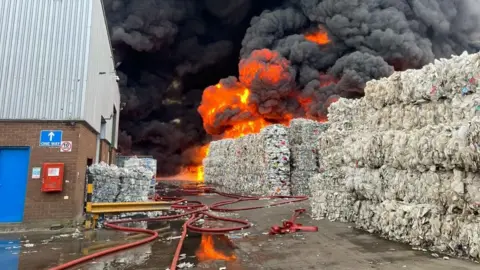 Steve Hubbard/BBC Bales of white-coloured waste stacked in five layers. Some of it is on fire and a huge black cloud of smoke is ascending from the fire. There is a warehouse building to the left with a brick ground floor and grey upper floor. There is a blue door. There is water on the yard between the building and the bales, with red hoses running across it.