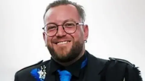 Police Scotland Smiling headshot of David Graham, who has a beard and is wearing glasses. He is wearing a black jacket, black shirt and a blue tie.