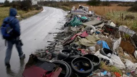 A man pictured walking on the footpath alongside huge amounts of rubbish which has been fly tipped.
