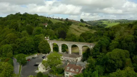 A drone photo of the mill in the countryside. A very scenic background with lots of trees and fields in the foreground. There is a viaduct in the middle of the picture with houses and a car park below. 