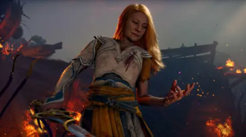 Sony Laufey is holding out her hand and looking at it, behind her things are on fire. She has long blond hair and silver armour on, she is holding a sword. 
