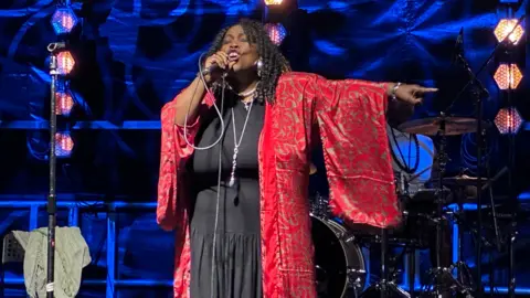 A woman in a black dress and a long red jacket is singing on a stage. Behind is a blue wall and a drum kit.