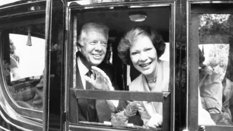 PA Media A black and white photo of Carter and his wife, Rosalynn, in a black carriage. Rosalynn is looking into the camera, smiling while Jimmy Carter looks to the side waving.