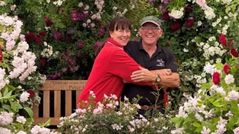 Tina and Ian Limmer embrace in a hug and smile at the cameras as they celebrate their win. They are surrounded by a display of roses with a brown bench in the background. Tina is wearing a red jumper and Ian a black polo neck. 