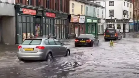Andrew Brammer Cars drive through flooding on a road, with waters lapping up to the side of shops and buildings at the side of the road.