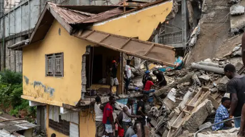     Prins Samura/EPA Afbeelding van een verwoest huis terwijl reddingswerkers en bewoners graven naar overlevenden in het puin van een ingestort gebouw in Freetown, Sierra Leone - maandag 16 september 2024.