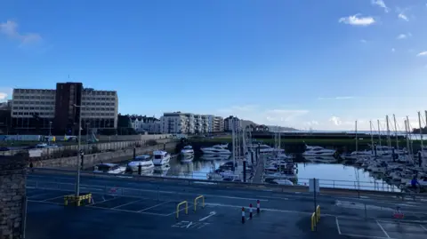 A marina. Boats are docked in the marina. The sky is blue. Tall buildings are on the left hand side.