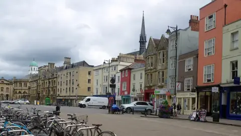 Stephen McKay A view of Broad Street in Oxford on an overcast day. Shops and University buildings such as The Bodleian Library can be seen. There are bike stands on the opposite side. Some vehicles are also parked on the street.