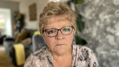 Tracy Canham stares at the camera. She has short blonde hair, dark eyes and thick-rimmed glasses. She is wearing a blouse covered in a snake skin print.