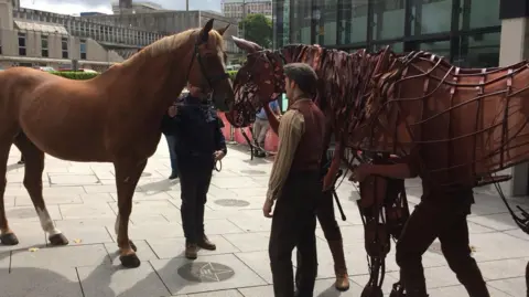The picture shows a real horse standing nose‑to‑nose with a full‑size horse puppet. The puppet is operated by visible puppeteers inside its frame, which is made of metal and shaped to look like the body of a horse. The scene takes place outdoors in a paved urban area, with modern buildings and glass walls in the background. 