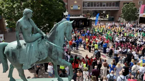 A shot from above of hundreds of people, mainly school children gathered in Broadgate main square in Coventry. A large statue of Lady Godiva on a horse is in the left of the picture and the crowd can be seen below. There is a stage in the top of the image where children are performing a musical about Lady Godiva.