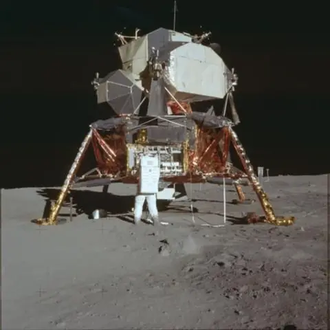 NASA The lunar lander stands squat and insect‑like on spindly golden legs, its foil‑wrapped descent stage glowing copper against the ash‑grey dust. Above, the pale, faceted cabin looks almost improvised – a fragile aluminium refuge dropped into a pristine desert. The harsh sunlight carves inky black shadows beneath the lander and across the pitted regolith, where bootprints and equipment puncture an otherwise untouched plain. 