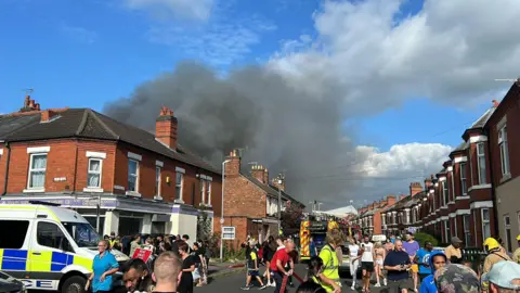 People milling around near emergency service vehicles with a plume of black smoke rising from behind nearby roofs