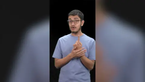 A man with a blue t shirt stood in front of a black background uses sign language.