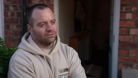 Carl Bumby, wearing a cream hooded jumper, stands in front of an open door with his arms crossed