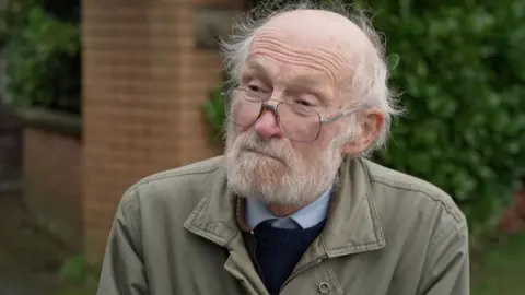 Bill wears glasses, a shirt, jumper and khaki jacket. He has a white grey beard.