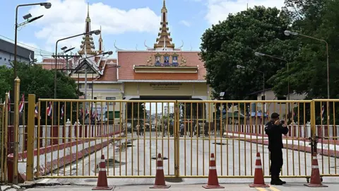 Shot of a checkpoint, with a roof built in the shape of a traditional temple. In front the checkpoint is a yellow gate and orange cones. A security official stands in front of the gate.