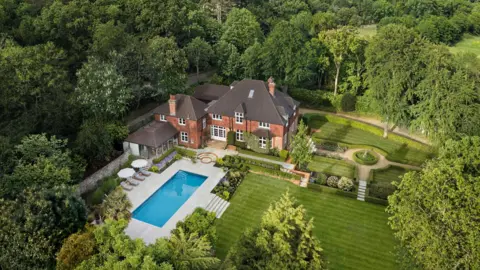 Omaze An aerial shot of a large house with a swimming pool set within a manicured garden and surrounded by trees