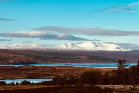 Fiona Morrill Snow-covered hills in the distance, with water and trees in the foreground.