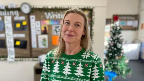 A person wearing a green Christmas jumper decorated with white reindeer and tree patterns stands in a classroom. Behind them is a festive display with a decorated Christmas tree and a noticeboard featuring writing materials and a clock. The room has bright lighting and a cheerful seasonal theme.