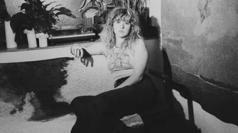 A black and white photo of a young woman with curly long hair and a full fringe sat on a sofa and looking into the camera with a serious expression. Her arm is resting on the side of the sofa and you can see plant pots on a shelf behind her. She is wearing a light vest top and dark trousers. 