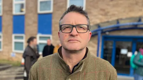 Tom Rainey in front of The King's Centre on Osney Mead. He is wearing glasses. There are other people behind him